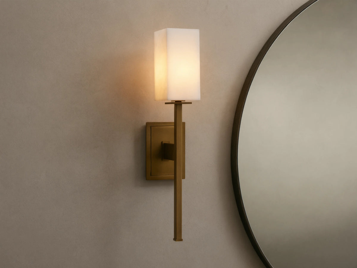 Cinda Sconce, a vertical brass finish wall light with a glowing rectangular stone shade, mounted on a tan wall next to a mirror.