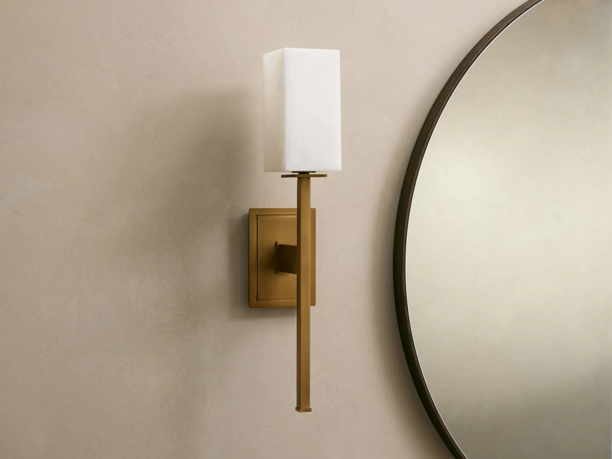 Cinda Sconce, a vertical brass finish wall light with a glowing rectangular stone shade, mounted on a tan wall next to a mirror.