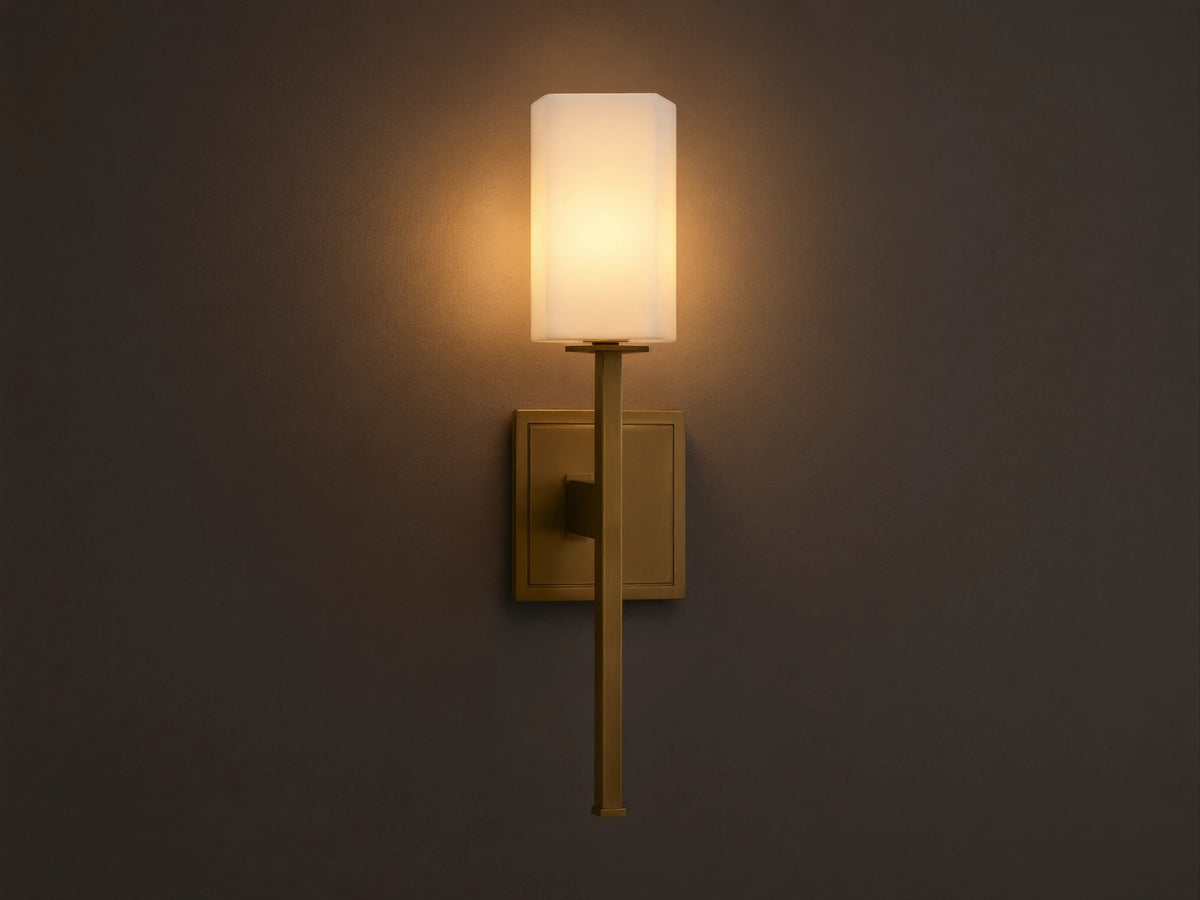 Cinda Sconce, a vertical brass finish wall light with a glowing rectangular stone shade, mounted on a tan wall next to a mirror.