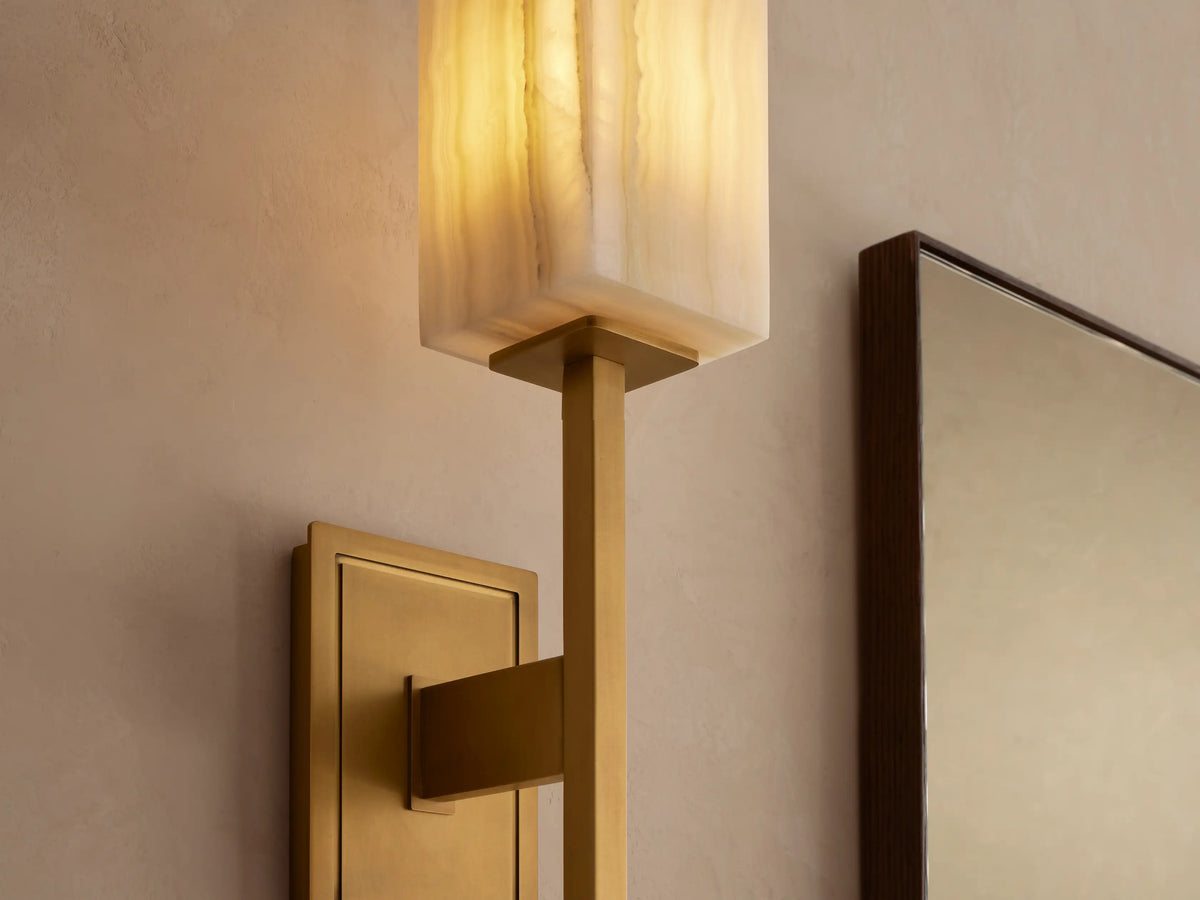 Cinda Sconce. Vertical brass-finished wall sconce with a rectangular, creamy, veined stone shade on a light wall next to a mirror.