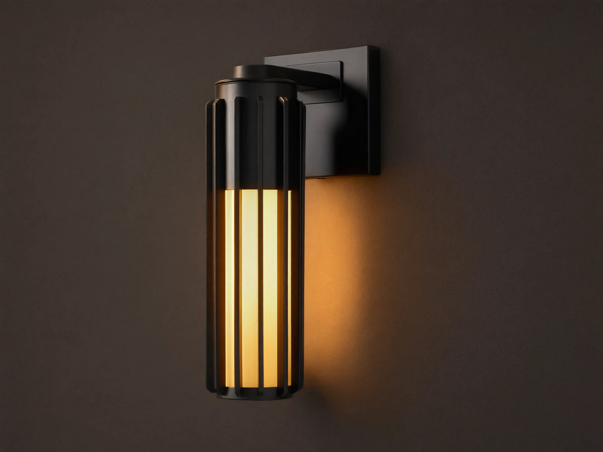 Matte black Gabbia Outdoor Sconce, cylindrical with vertical slats, illuminated warm milk glass, mounted on a beige wall.