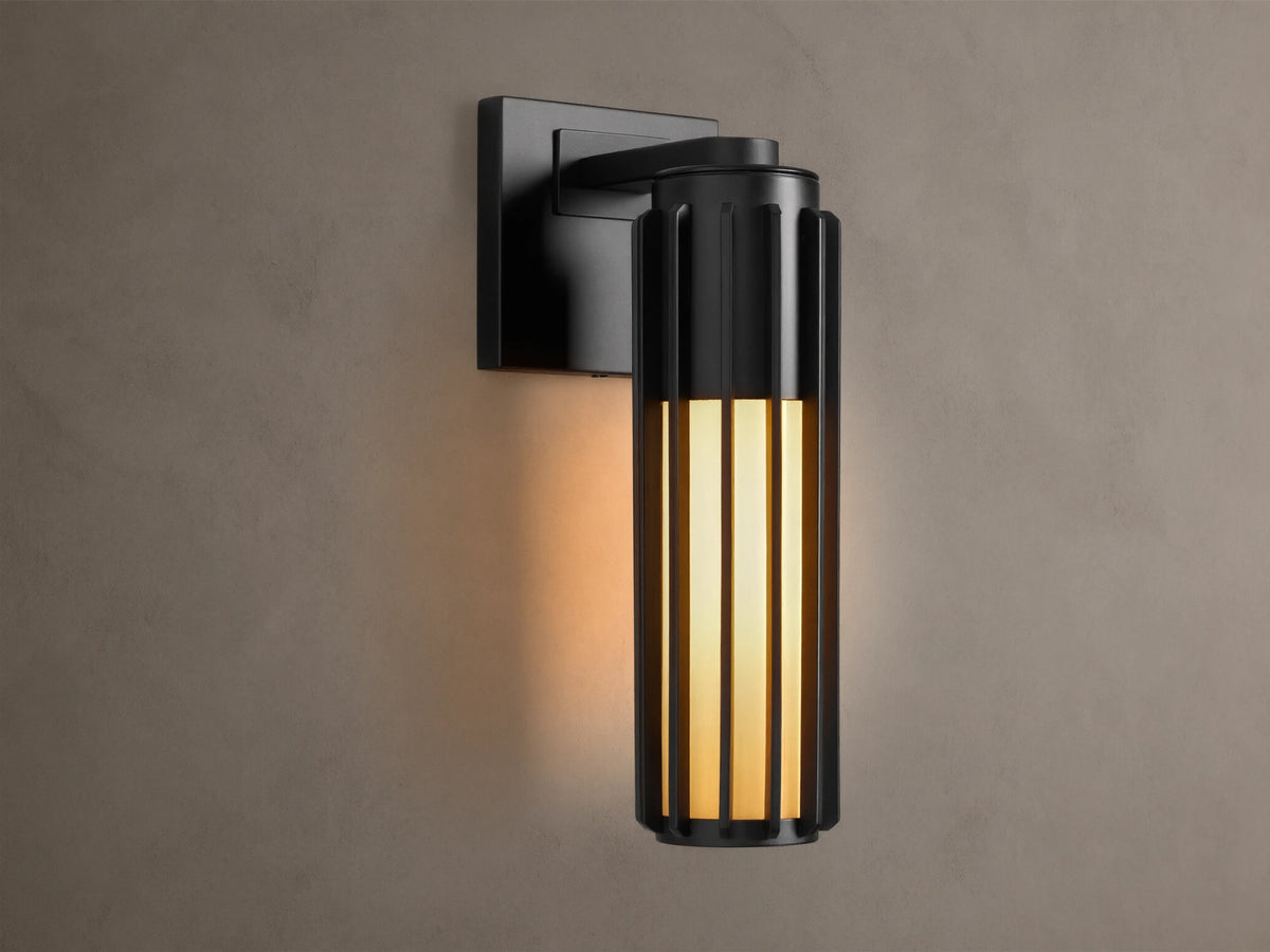 Matte black Gabbia Outdoor Sconce, cylindrical with vertical slats, illuminated warm milk glass, mounted on a beige wall.