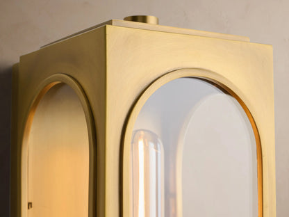 Vertical brass Palladian Outdoor Sconce with arched glass windows, showing a long filament bulb, mounted on a tan wall.