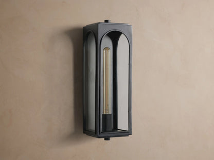 Black Palladian Outdoor Sconce, vertical, with arched glass panels and a warm glowing filament bulb, mounted on a tan wall.