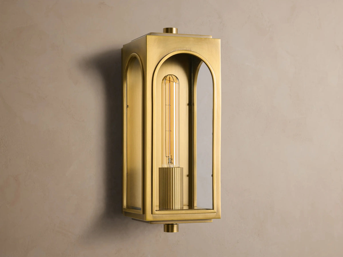 Palladian Outdoor Sconce: Polished brass rectangular fixture with arched glass, bright tubular bulb, mounted on a grey wall.
