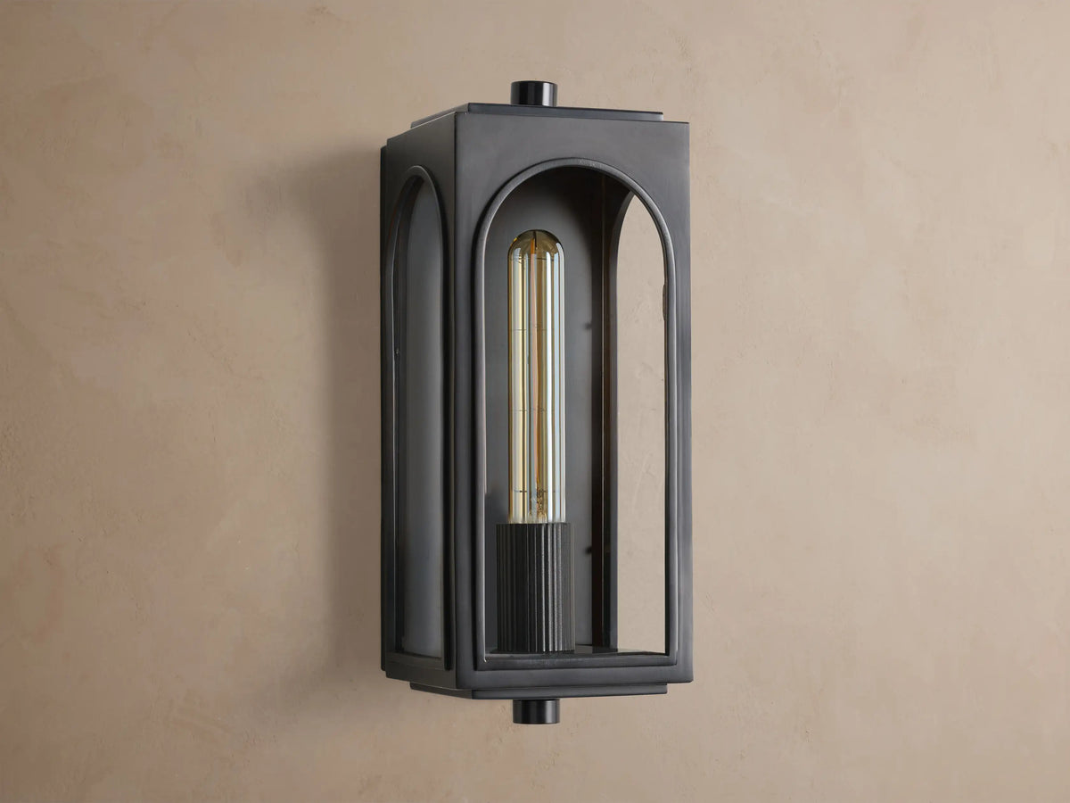 Dark plated brass Palladian Outdoor Sconce, rectangular with arched glass, warm glowing filament bulb on a brown wall.