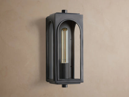 Dark plated brass Palladian Outdoor Sconce, rectangular with arched glass, warm glowing filament bulb on a brown wall.