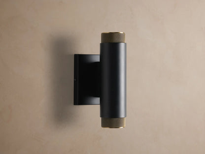 Matte black cylindrical Spotlight Outdoor Sconce with brass knurled end caps, mounted vertically on a light brown wall.