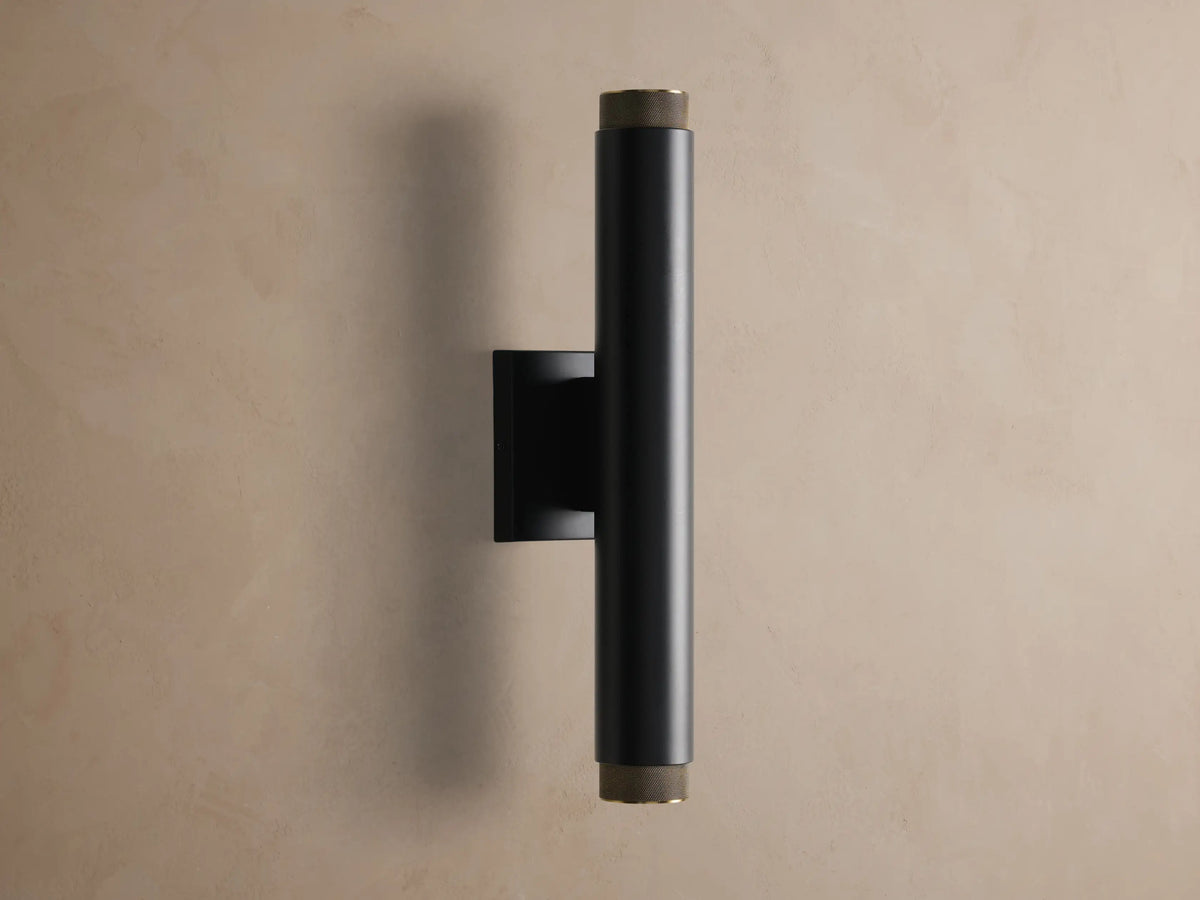Vertical matte black cylindrical Spotlight Outdoor Sconce with brass knurled ends, mounted on a textured beige wall.