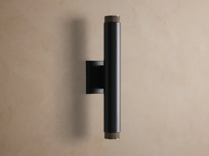Vertical matte black cylindrical Spotlight Outdoor Sconce with brass knurled ends, mounted on a textured beige wall.