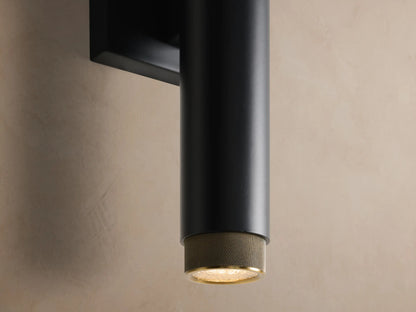 Matte black cylindrical Spotlight Outdoor Sconce with brass knurled end caps, mounted vertically on a light brown wall.