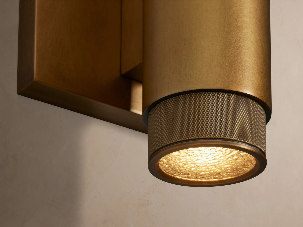 Brass Spotlight Outdoor Sconce, cylindrical, with knurled ends, mounted vertically on a light wall. Warm light shines from behind.