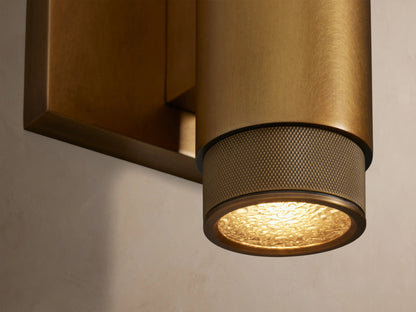 Brass Spotlight Outdoor Sconce, cylindrical, with knurled ends, mounted vertically on a light wall. Warm light shines from behind.