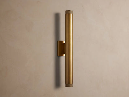 Vertical brass spotlight outdoor sconce with cylindrical body and knurled end caps, mounted on a neutral wall.