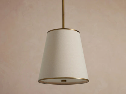 Adria Pendant with a light fabric conical shade, brass trim, hanging and emitting a warm glow against a textured wall.