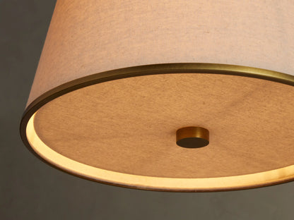 Adria Pendant with conical cream fabric shade and brass trim, hanging against a soft, textured beige wall.