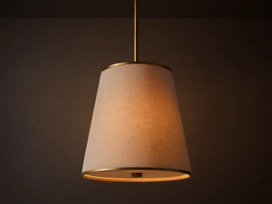 Adria Pendant with a light fabric conical shade, brass trim, hanging and emitting a warm glow against a textured wall.