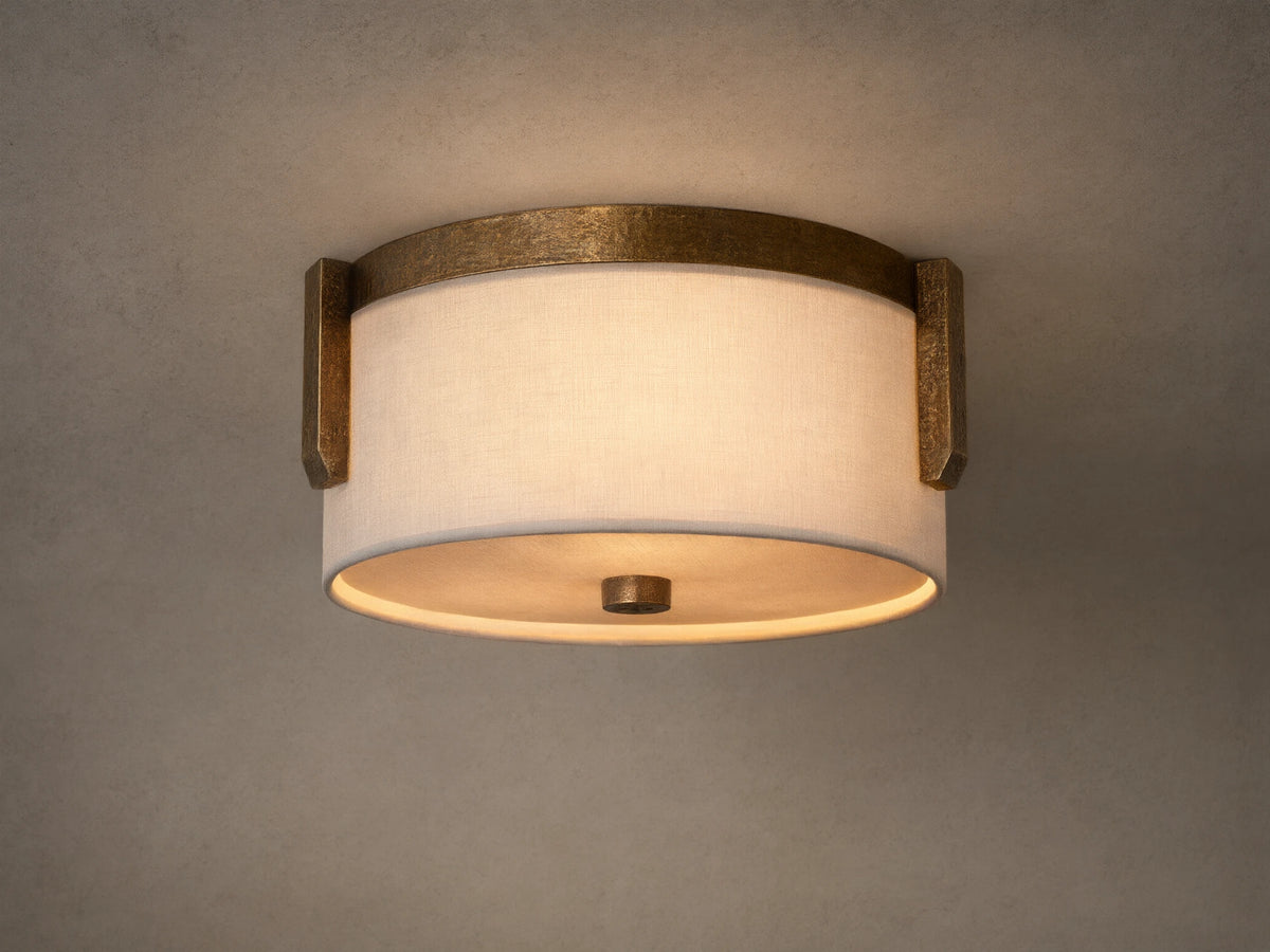 Emmet Flush Mount. Illuminated round white linen shade framed by textured antiqued brass, mounted on a light-colored ceiling.