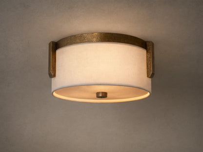 Emmet Flush Mount. Illuminated round white linen shade framed by textured antiqued brass, mounted on a light-colored ceiling.