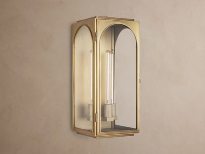 Lit brass Palladian Outdoor Sconce with arched glass panels, showing two warm filament bulbs, mounted on a textured gray wall.