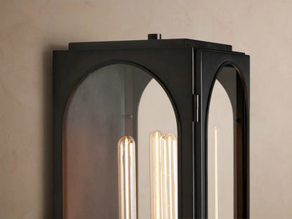 Vertical Palladian Outdoor Sconce, dark metal with two arched clear glass panels, showcasing a glowing filament bulb, on a textured wall.