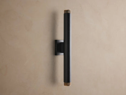 Black Spotlight Outdoor Sconce, vertically mounted, with brass knurled ends, illuminating a textured tan wall.