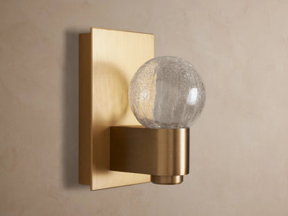 Casen Sconce with satin brass rectangular backplate and spherical crackle glass globe glowing warmly on a wall.