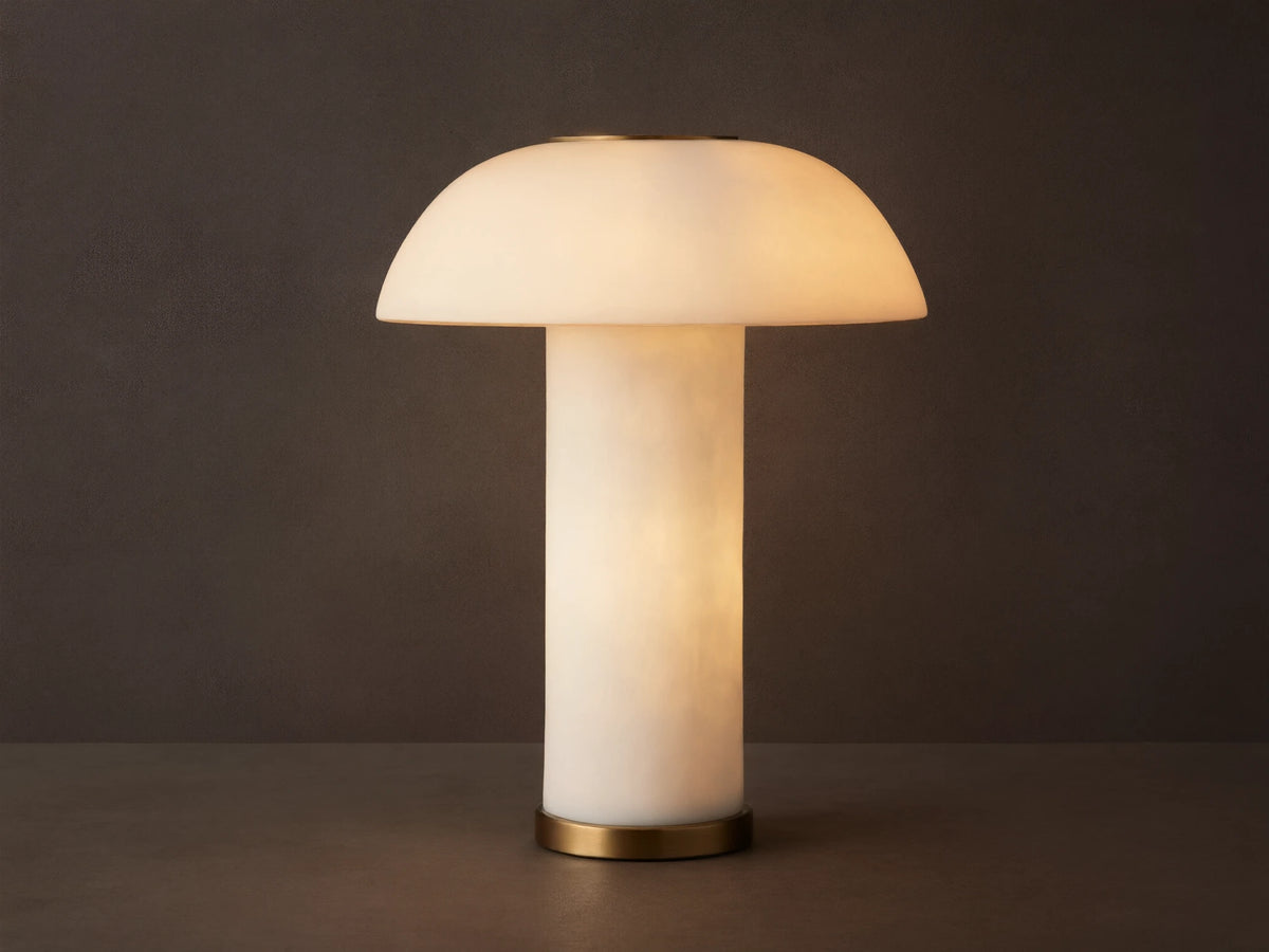 Lit Tramonto Table Lamp, mushroom-shaped, translucent cloudstone resin with brass accents, glowing on a rustic wooden table.