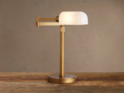 Zayd Swing Arm Table Lamp with brass finish and illuminated white opal glass shade, on a wooden table.