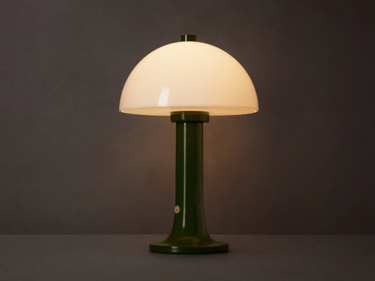 Jocy Portable Table Lamp with a dark green lacquered base and a domed white opal shade, illuminated on a wooden table.