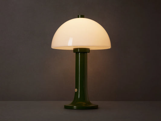 Jocy Portable Table Lamp with a dark green lacquered base and a domed white opal shade, illuminated on a wooden table.