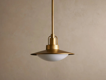 A brass pendant light with a broad, flat circular shade hangs from the ceiling. It emits a warm glow against a neutral backdrop.