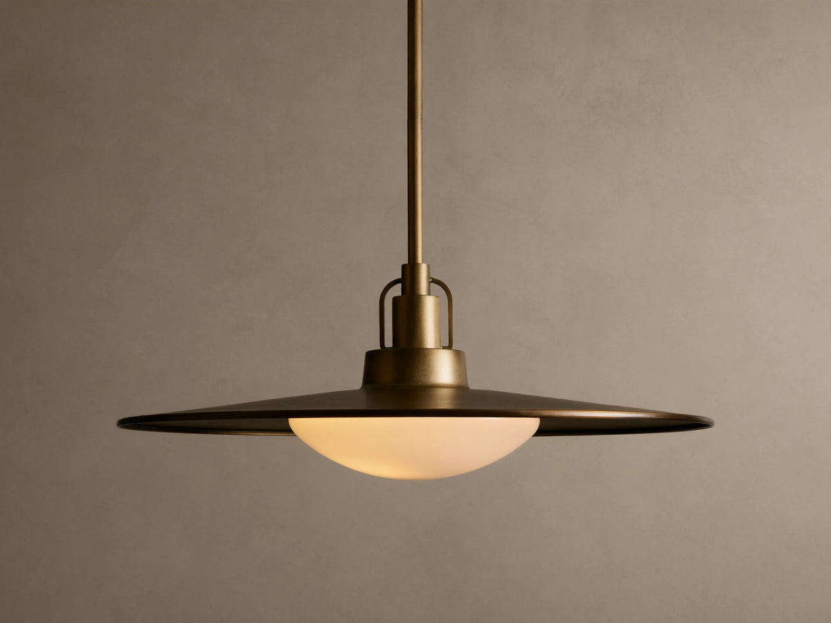 Brass pendant light with a wide, flat shade and a warm glowing bulb. It hangs from a rod against a beige textured wall.