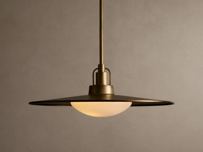 Brass pendant light with a wide, flat shade and a warm glowing bulb. It hangs from a rod against a beige textured wall.