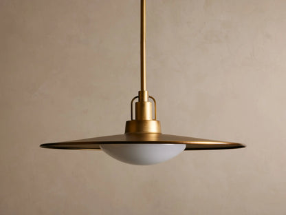 Brass pendant light with a wide, flat shade and a warm glowing bulb. It hangs from a rod against a beige textured wall.