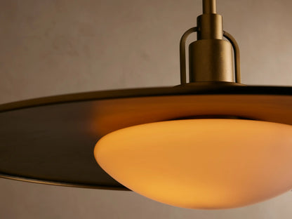 A brass pendant light with a wide, flat shade and frosted white bulb hangs centered against a beige wall.