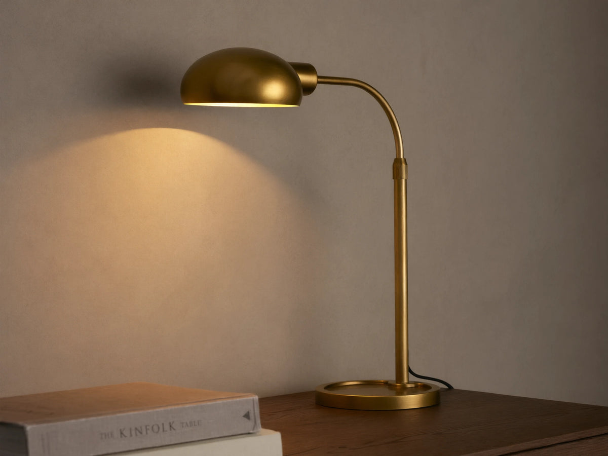 Brass-finished Bergamo Task Table Lamp with a domed shade and curved arm, emitting warm light onto a concrete wall above books on a desk.