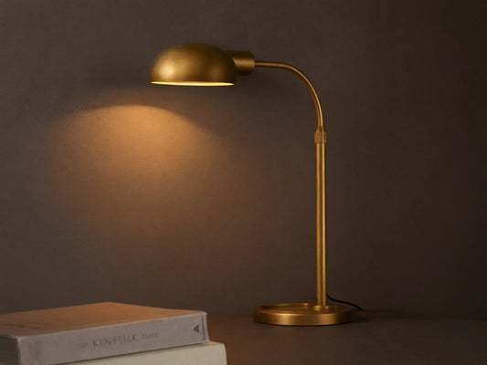 Brass-finished Bergamo Task Table Lamp with a domed shade and curved arm, emitting warm light onto a concrete wall above books on a desk.