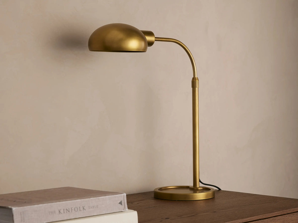 Brass-finished Bergamo Task Table Lamp with a domed shade and curved arm, emitting warm light onto a concrete wall above books on a desk.