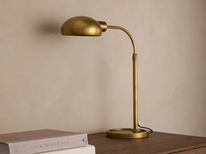 Brass-finished Bergamo Task Table Lamp with a domed shade and curved arm, emitting warm light onto a concrete wall above books on a desk.