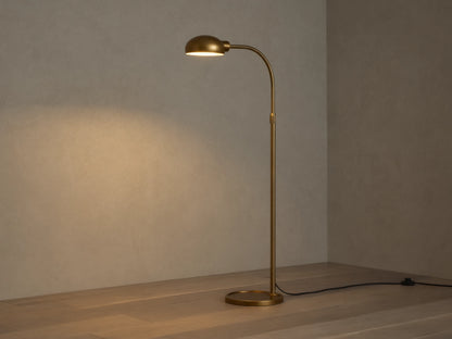Sleek brass Bergamo Task Floor Lamp with illuminated, pivoting shade, standing on wood floor against a textured wall.