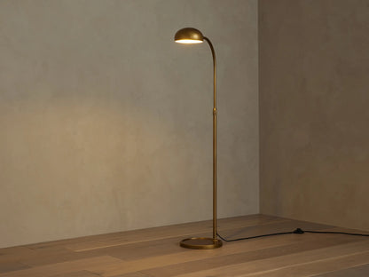 Brass Bergamo Task Floor Lamp with curved arm, round shade, and base on wood floor. Black cord with dimmer switch.
