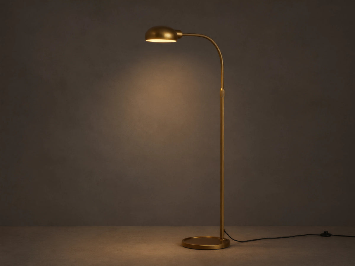 Sleek brass Bergamo Task Floor Lamp with illuminated, pivoting shade, standing on wood floor against a textured wall.