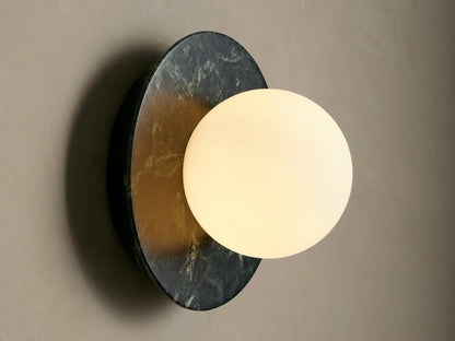 Corinna Sconce: Round dark green marble backplate with white veins, supporting a spherical milk glass globe on a beige wall.