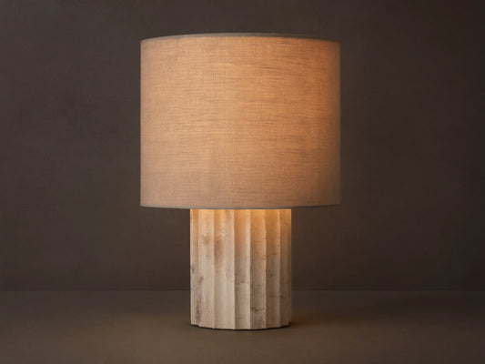 A beige table lamp with a cylindrical fabric shade emits warm light. It stands on a textured, fluted stone base, placed on a wooden table.