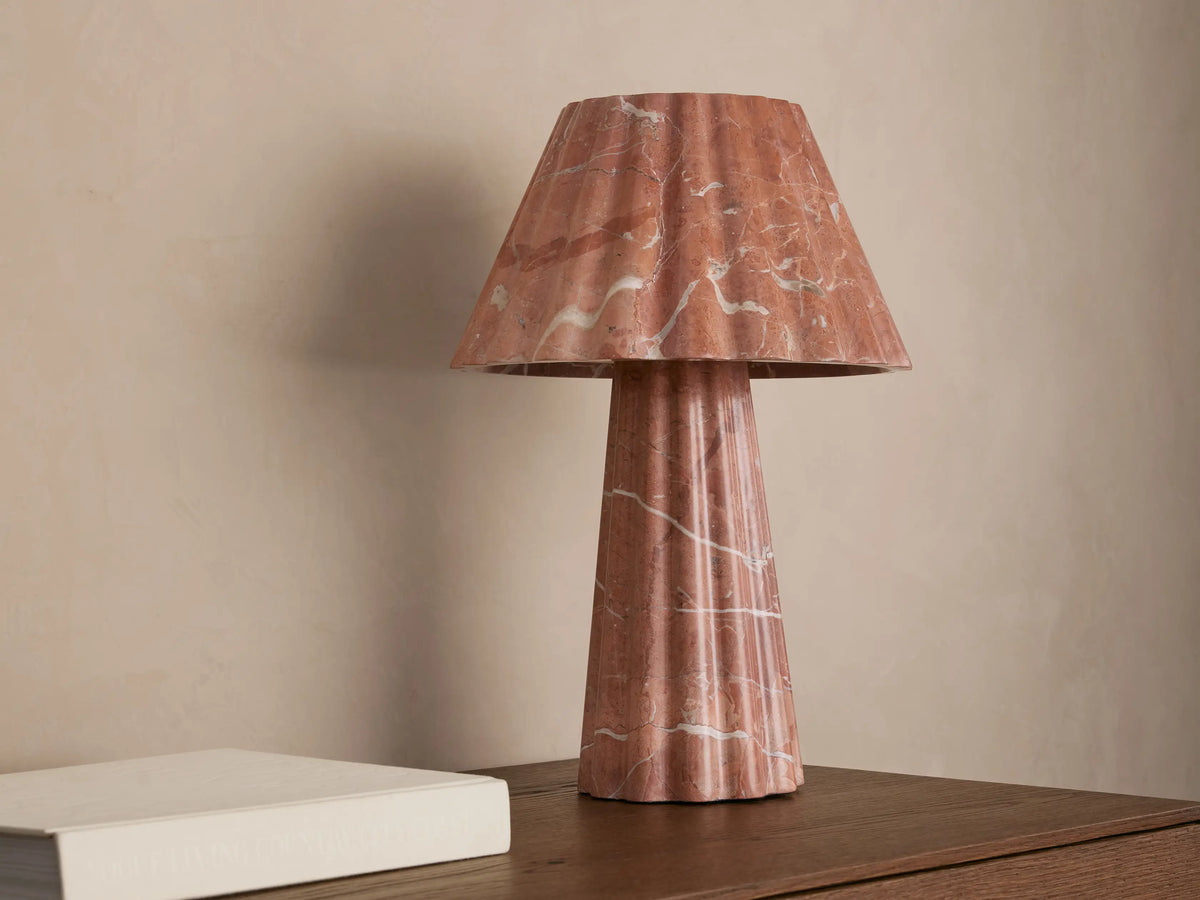 A pink marble table lamp with a fluted base and conical shade casts a warm light on a wooden surface next to a closed book.