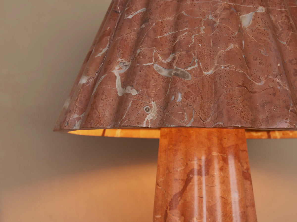 A pink marble table lamp with a cone-shaped shade and base featuring white veining, set on a wooden surface beside a closed book.