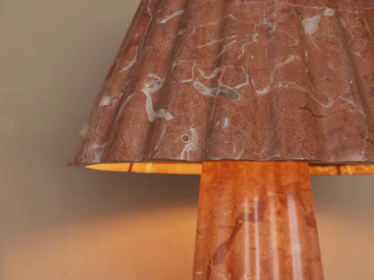 A pink marble table lamp with a cone-shaped shade and base featuring white veining, set on a wooden surface beside a closed book.