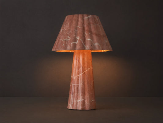 A pink marble table lamp with a fluted base and conical shade casts a warm light on a wooden surface next to a closed book.