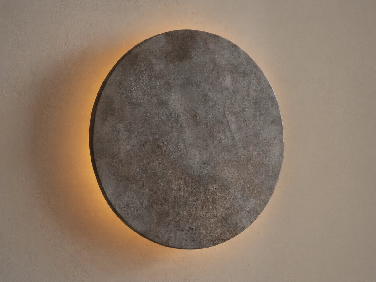 Odette Outdoor Sconce: Oval dark grey natural stone sconce on a beige wall, emitting warm golden LED light from behind.